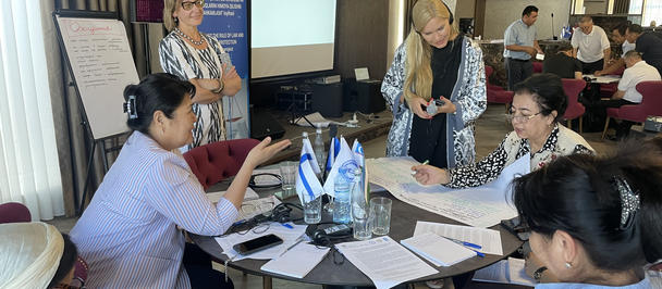 Group of people engaged in a discussion around a table with documents and a presentation screen.